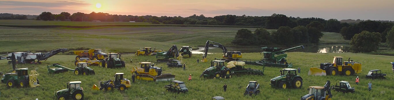 ಒಂದು ದೊಡ್ಡ ಹೊಲದಲ್ಲಿ ಜನರು ಅನೇಕ ರೀತಿಯ John Deere ಸಲಕರಣೆಗಳ ಮಧ್ಯೆ ನಡೆಯುತ್ತಿದ್ದಾರೆ.